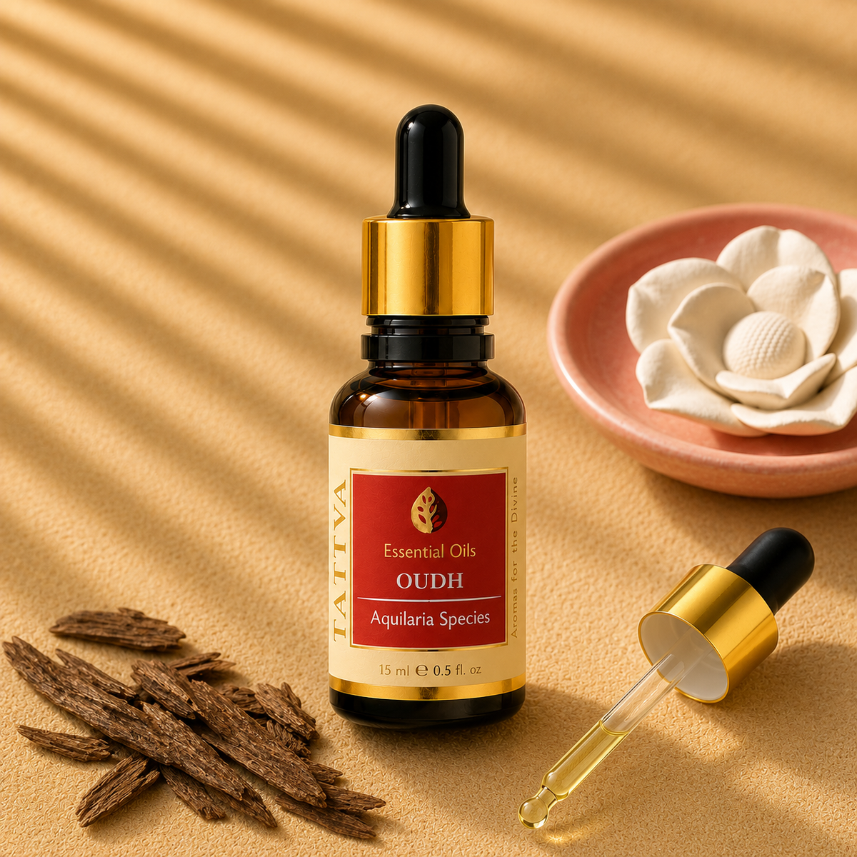 A small amber glass dropper bottle labeled 'OUDH' and 'Essential Oils' stands on a warm beige surface with shadow bands across it. A gold-and-black dropper lies nearby, with dried brown bark pieces at left and a pink dish with a white flower at right.