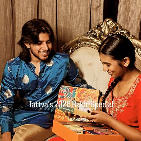 A young man in a blue patterned kurta and a woman in a red dress sitting on an ornate sofa, smiling as they open a colorful gift box from Tattva Aromas. The text 'Tattva's 2026 Rakhi Special' is overlaid on the center.